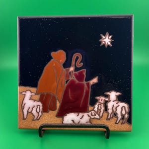 May include: A square ceramic tile featuring a nativity scene. Two figures in robes stand before a star, with sheep in the foreground. The background is dark blue with white speckles, and the figures are in shades of brown and red.