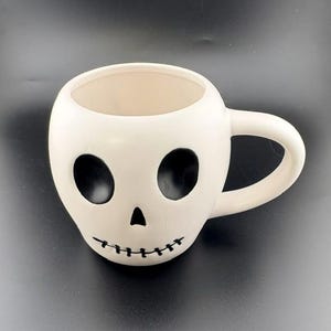 May include: A white ceramic mug shaped like a human skull. The mug features two large, hollow black eye sockets, a small triangular black nose cavity, and a thin black line representing a stitched mouth. It has a curved handle on the right side.
