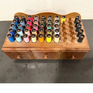 May include: A wooden sewing box with three drawers and a top section for thread spools. The box is filled with colorful thread spools in various colors, including blue, pink, yellow, and black. The box has a dark brown finish.