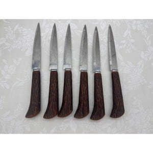 May include: Set of six vintage steak knives. Each knife has a stainless steel blade and a dark brown, textured handle. The handles have a curved, ergonomic design. Knives are arranged on a white floral patterned surface.