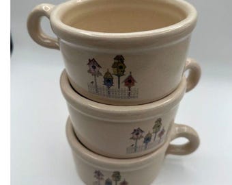 May include: A stack of three cream-colored ceramic mugs, each featuring a repeating graphic print of colorful birdhouses on posts behind a white picket fence. The mugs have a smooth, rounded shape with a single handle on the side.