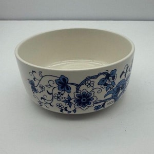 May include: Off-white ceramic bowl with a blue floral pattern around the rim. The bowl has a simple, rounded shape and a smooth, glossy finish. The floral design features stylized flowers and leaves.