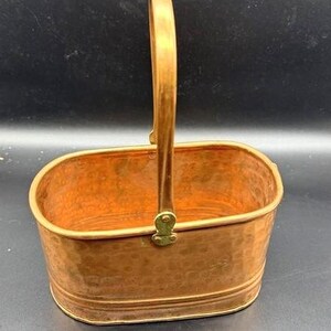 May include: A hammered copper basket with a brass handle. The basket is oval shaped and has a textured surface.