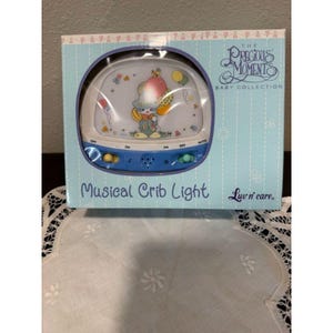 May include: A baby crib light in a box with the text "The Precious Moments Baby Collection" and "Musical Crib Light". The light has a blue base with buttons and a picture of a cartoon character holding balloons. The box is light blue.