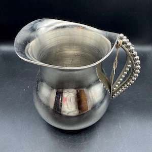 May include: A silver-colored pitcher with a rounded body and a flared spout. The handle is adorned with a beaded design. The pitcher reflects the surrounding environment, suggesting a polished surface. A classic piece for serving beverages.