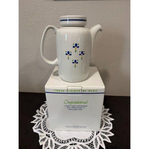 May include: White ceramic coffee pot with blue floral design and a blue stripe around the lid. The pot is on top of a white box with the text "FRESH FLOWER BY AVON" and "Congratulations!" printed on it.