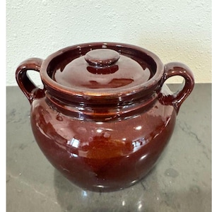 May include: A dark brown glazed ceramic bean pot with a matching lid and two side handles. The pot features a rounded body and a smooth, glossy finish that reflects light, showcasing a deep, uniform color throughout the entire exterior surface.