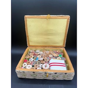 May include: A rectangular sewing box with a woven exterior and a hinged lid, featuring a pale yellow interior lining. The box is filled with numerous small wooden spools of thread in various colors and a white rectangular case with red stripes.