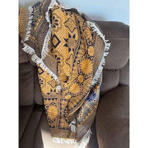 May include: A round, patterned throw blanket with a yellow and navy blue design. The blanket features geometric and floral motifs, and is trimmed with white fringe. The blanket is draped over a brown upholstered chair.