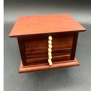 May include: A rectangular wooden box with a dark reddish-brown finish, featuring a flat top and a front panel composed of horizontal slats. A vertical string of small, round, off-white beads is attached to the center of the front panel.