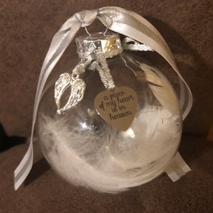 May include: A clear glass ornament with white feathers inside. It has a silver ribbon and a silver charm with angel wings. The charm has a heart-shaped tag that reads "a piece of my heart is in heaven".