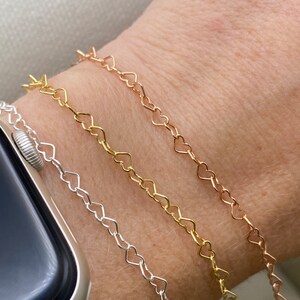 May include: Three heart-shaped chain bracelets in silver, gold, and rose gold. The bracelets are layered on a person's wrist.