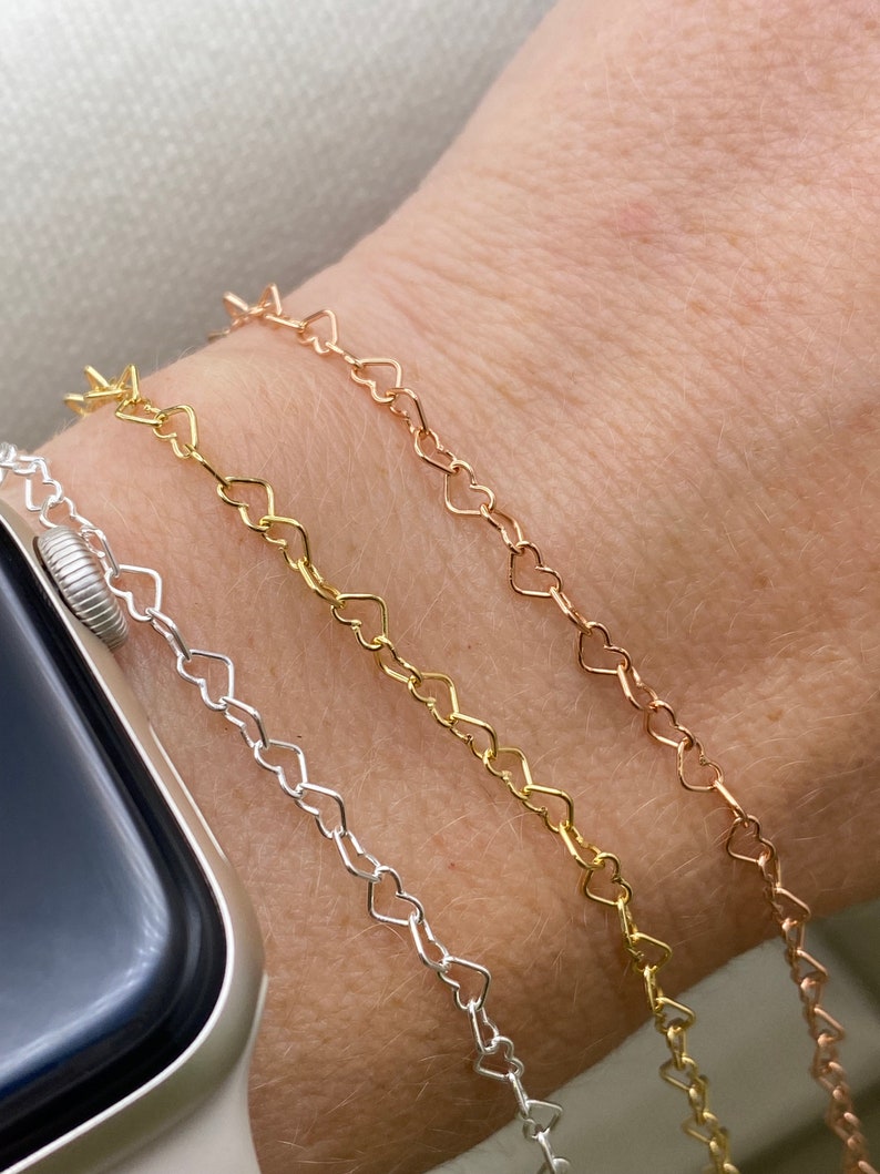 May include: Three heart-shaped chain bracelets in silver, gold, and rose gold. The bracelets are layered on a person's wrist.