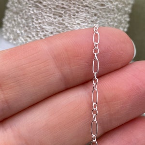 May include: Close-up of a silver-colored chain with an alternating oval and round link design. The chain is held between fingers, with a roll of chain visible in the background. This jewelry chain is ideal for crafting necklaces and bracelets.