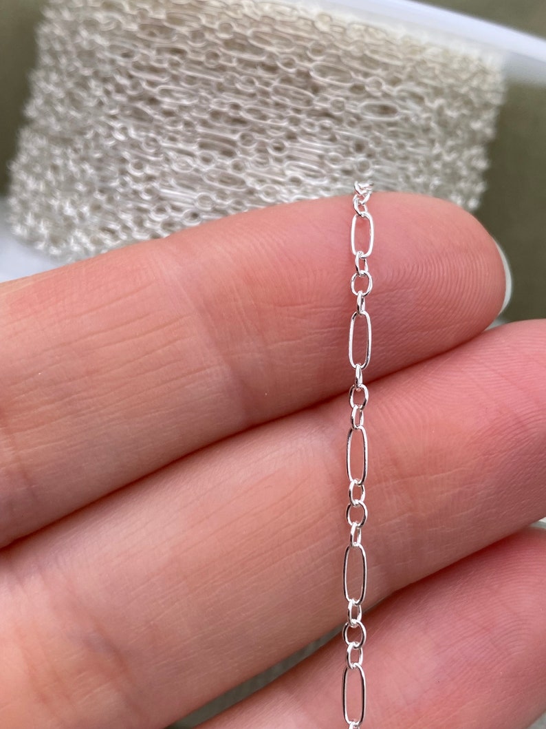 May include: Close-up of a silver-colored chain with an alternating oval and round link design. The chain is held between fingers, with a roll of chain visible in the background. This jewelry chain is ideal for crafting necklaces and bracelets.