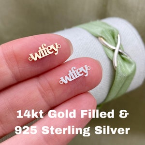 May include: Two small, delicate charms, one gold-colored and one silver-colored, each spelling out the word "wifey". The charms have small loops on each end. The text "14kt Gold Filled & 925 Sterling Silver" is visible below the charms.