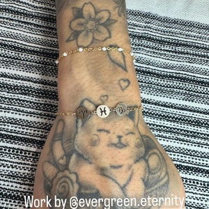 May include: Two gold bracelets on a tattooed wrist. The top bracelet has white beads. The bottom bracelet has three gold charms with zodiac signs: Aries, Pisces, and Virgo. The text "Work by @evergreen.eternity" is at the bottom of the image.