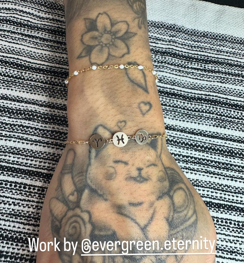 May include: Two gold bracelets on a tattooed wrist. The top bracelet has white beads. The bottom bracelet has three gold charms with zodiac signs: Aries, Pisces, and Virgo. The text "Work by @evergreen.eternity" is at the bottom of the image.