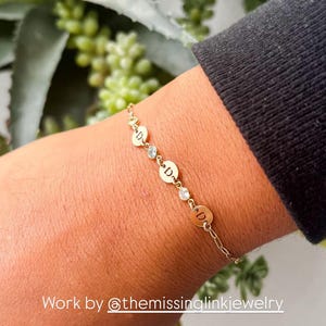 May include: Gold chain bracelet with small round charms featuring the letter "D" and a clear rhinestone between each charm. The bracelet is on a person's wrist. Work by @themissinglinkjewelry