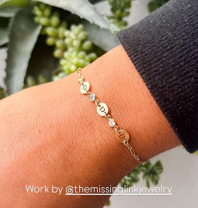 May include: Gold chain bracelet with small round charms featuring the letter "D" and a clear rhinestone between each charm. The bracelet is on a person's wrist. Work by @themissinglinkjewelry