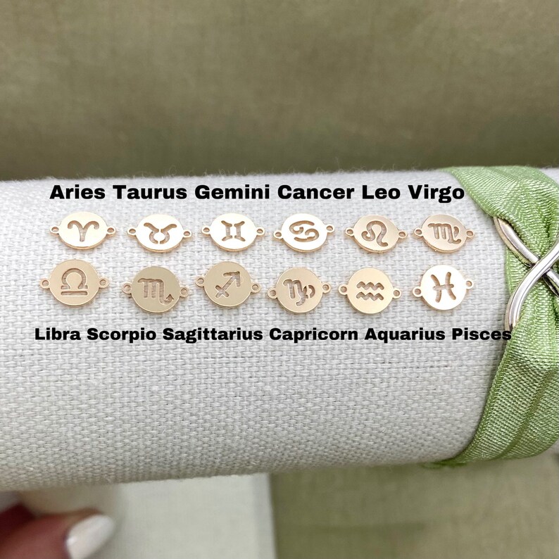 May include: Gold-toned zodiac charms with the astrological symbols for Aries, Taurus, Gemini, Cancer, Leo, Virgo, Libra, Scorpio, Sagittarius, Capricorn, Aquarius, and Pisces. The charms are arranged in two rows on a light beige fabric background.