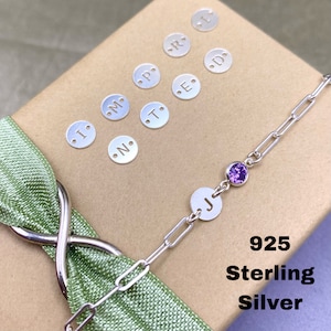 May include: A silver chain bracelet with a small purple gemstone and a charm with the letter "J" on it. The bracelet is on a brown surface with a green ribbon and silver letter charms.