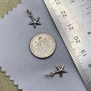 May include: Two silver starfish charms, one with a loop for attaching to a necklace or bracelet, the other with a jump ring. The charms are shown next to a US dime for size comparison.