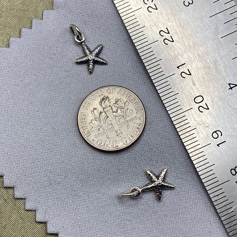May include: Two silver starfish charms, one with a loop for attaching to a necklace or bracelet, the other with a jump ring. The charms are shown next to a US dime for size comparison.