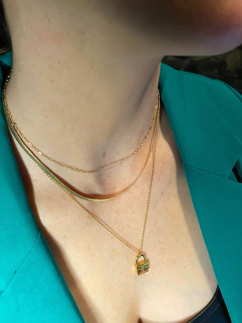 May include: A gold-colored layered necklace set. The necklace features three chains: a herringbone chain, a delicate chain, and a pendant necklace with a small padlock charm. The model is wearing a teal blazer.