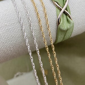 May include: Three chains, one gold and two silver, are displayed on a fabric surface. The chains are all the same style, a twisted rope chain.