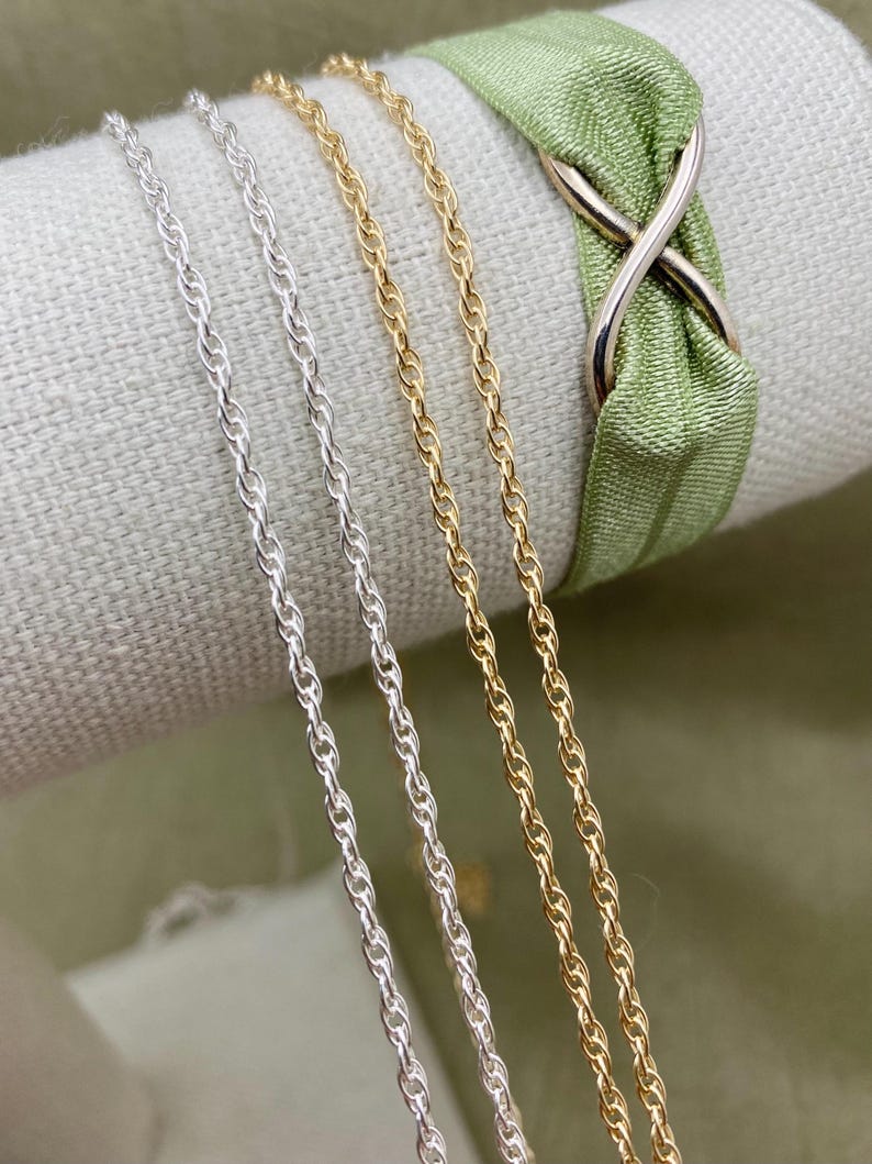 May include: Three chains, one gold and two silver, are displayed on a fabric surface. The chains are all the same style, a twisted rope chain.