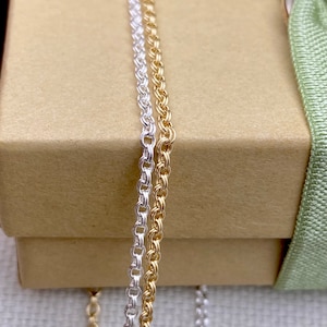May include: Two chains, one silver and one gold, are displayed on a brown box. The chains are both twisted and have a delicate, intricate design.