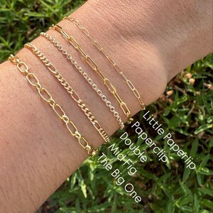 May include: Five gold chain bracelets stacked on a person's wrist. The bracelets are labelled from top to bottom: Little Paperlip, Paperclip, Double, Mid Fig, The Big One.
