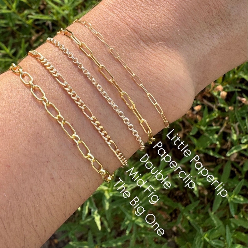 May include: Five gold chain bracelets stacked on a person's wrist. The bracelets are labelled from top to bottom: Little Paperlip, Paperclip, Double, Mid Fig, The Big One.
