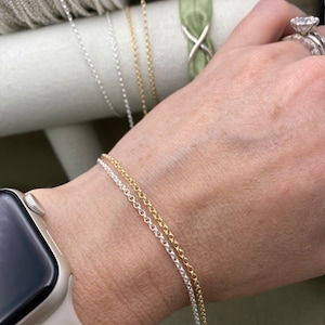 May include: Two silver and gold chains on a person's wrist. The chains are delicate and have a simple design.