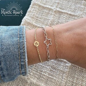 May include: Three gold and silver chain bracelets with charms. The charms are a star, an Om symbol, and a small circle. The bracelets are stacked on a person's wrist.