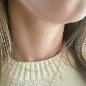 May include: Three delicate gold necklaces layered together. The top chain is a small oval link design, the middle chain is a paperclip style, and the bottom chain is a simple pendant necklace. The necklaces are worn over a cream-colored sweater.