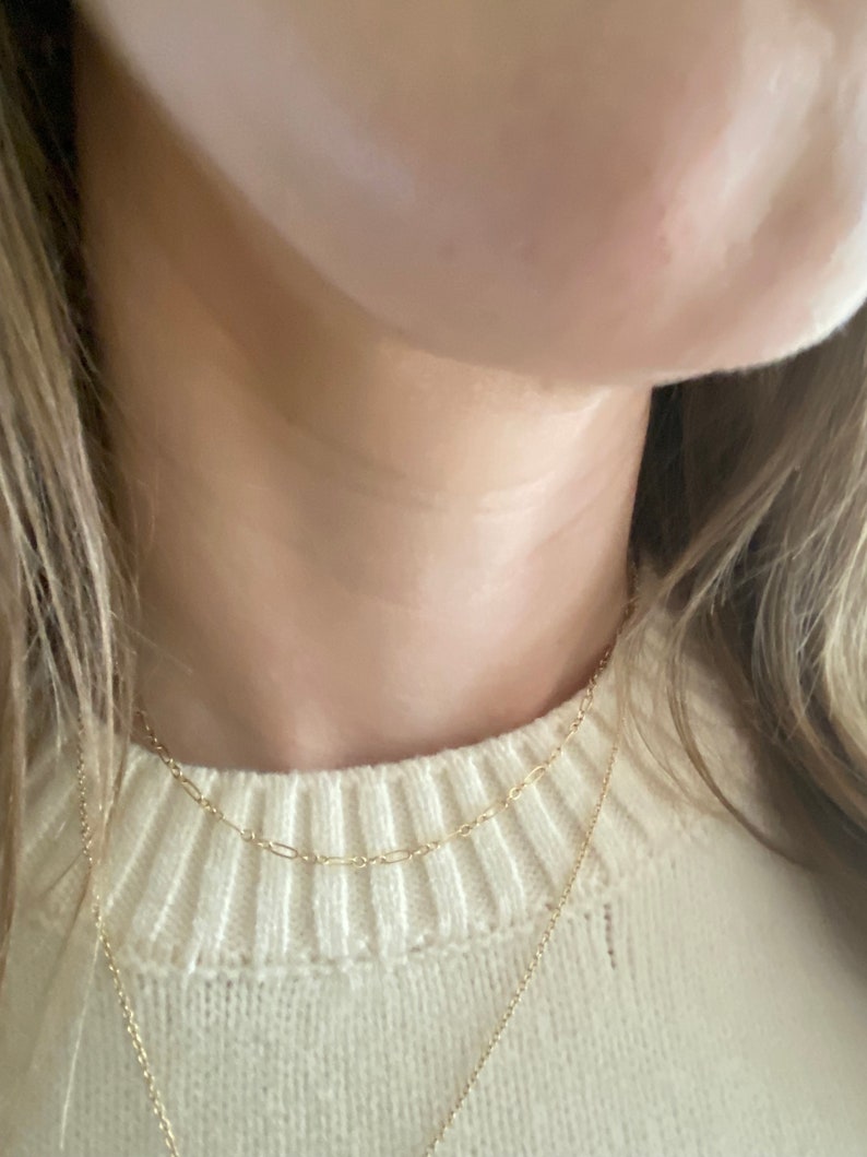 May include: Three delicate gold necklaces layered together. The top chain is a small oval link design, the middle chain is a paperclip style, and the bottom chain is a simple pendant necklace. The necklaces are worn over a cream-colored sweater.