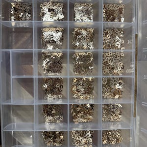 Pode incluir: Uma caixa de armazenamento de pl&aacute;stico transparente com 18 compartimentos cheios de contas de letras de metal douradas. As contas est&atilde;o dispostas em um padr&atilde;o de grade.
