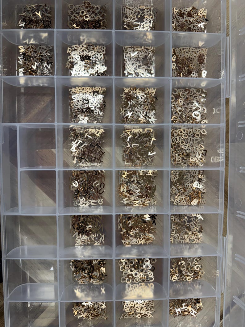 Pode incluir: Uma caixa de armazenamento de pl&aacute;stico transparente com 18 compartimentos cheios de contas de letras de metal douradas. As contas est&atilde;o dispostas em um padr&atilde;o de grade.