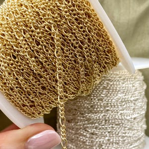 May include: Close-up of two spools of jewelry chain. One spool is gold-colored, and the other is silver-colored. The gold chain is in focus, with a delicate, linked design. The silver chain is slightly out of focus in the background. The chains are on white spools.