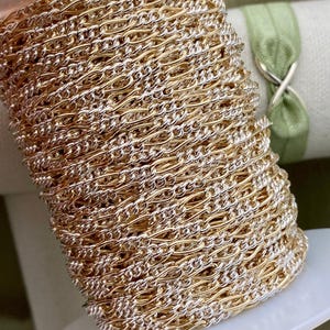 May include: A spool of gold and silver-toned curb chain. The chain is tightly wound on a white plastic spool. The chain is a mix of gold and silver links, creating a two-tone effect. The background includes a green ribbon and a white fabric.