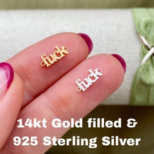 May include: Two small charms, one gold-coloured and one silver-coloured, each spelling the word "fuck" in a cursive font. The text "14kt Gold filled & 925 Sterling Silver" is displayed below the charms.