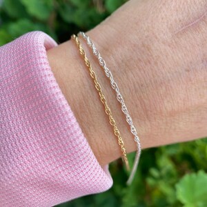 May include: Two bracelets on a wrist, one gold and one silver. Both bracelets are rope chain style.
