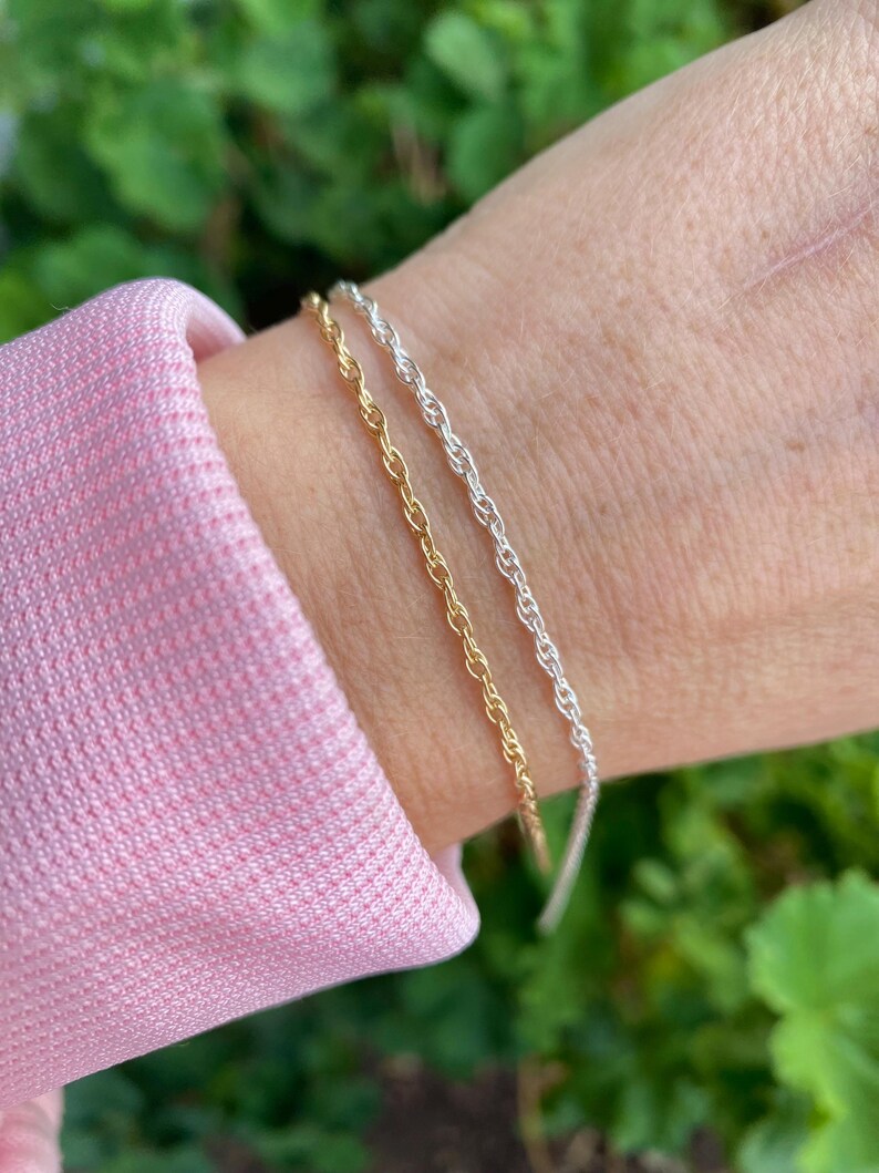May include: Two bracelets on a wrist, one gold and one silver. Both bracelets are rope chain style.