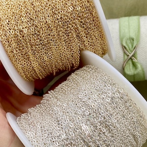 Könnte beinhalten: Nahaufnahme von zwei Spulen mit filigraner Kette. Eine Spule ist goldfarben, die andere silberfarben. Die Ketten bestehen aus kleinen, miteinander verbundenen Gliedern. Die Spulen sind weiß und vor einem sanften grünen Hintergrund platziert.