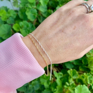 May include: Two gold and silver rope chain bracelets on a wrist. The bracelets are layered on top of each other.