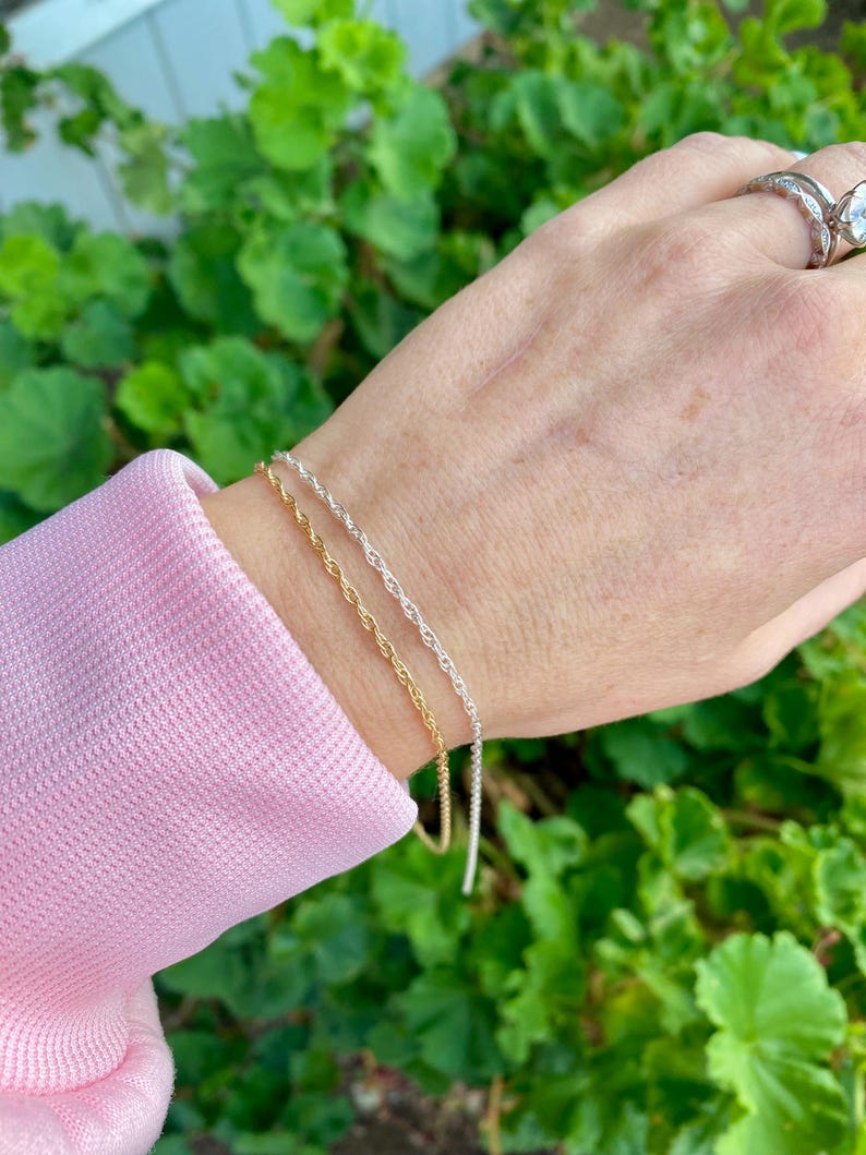 May include: Two gold and silver rope chain bracelets on a wrist. The bracelets are layered on top of each other.