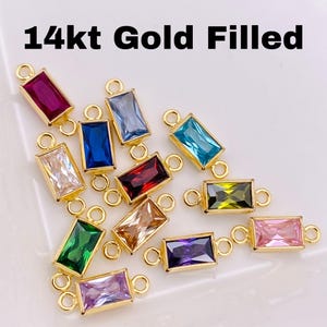 May include: Gold-filled rectangular charms with various colored rectangular gemstones. The charms have a gold frame and small loops on each end. The text "14kt Gold Filled" is at the top of the image. The gemstones are in shades of red, blue, green, and pink.