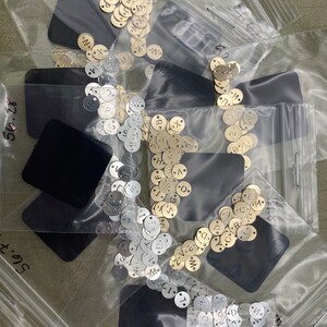 May include: Three clear plastic bags filled with small round metal discs. The discs are silver, gold, and brass and have letters stamped on them. The bags are on a green surface.